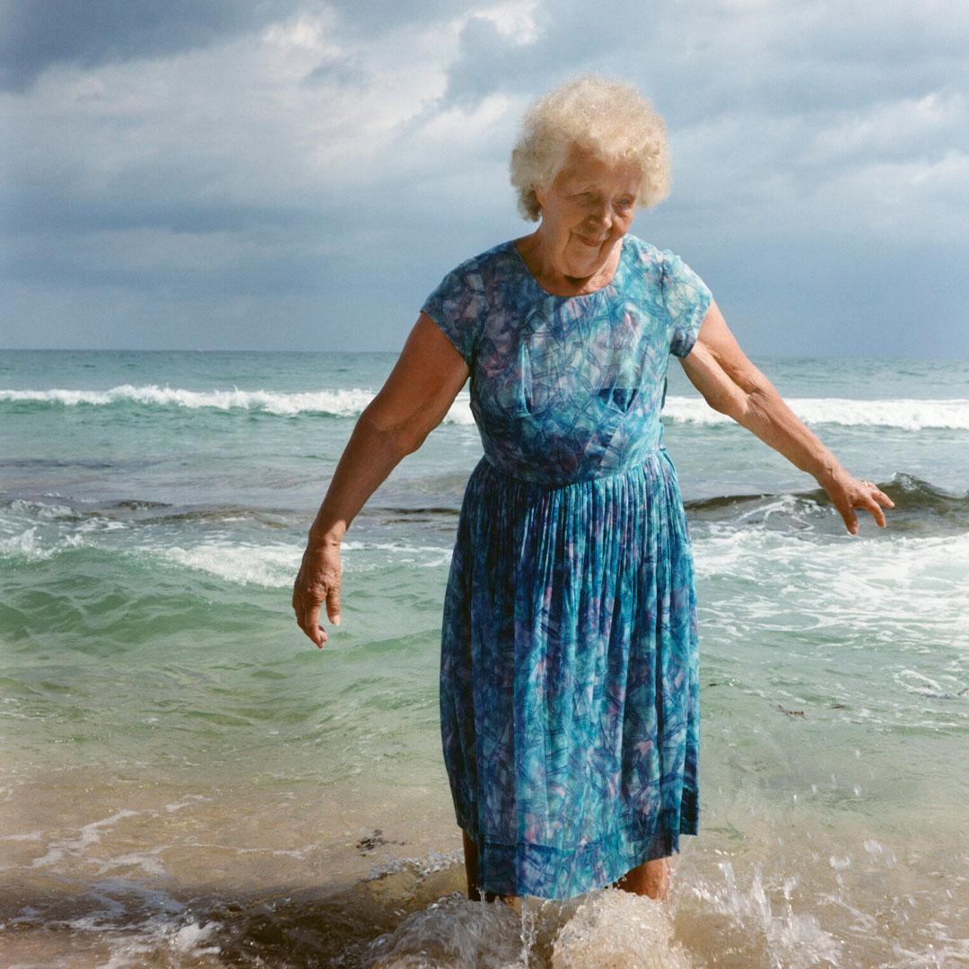 En äldre kvinna med kort vitt hår och blå klänning står med fötterna i havet vid en strandkant